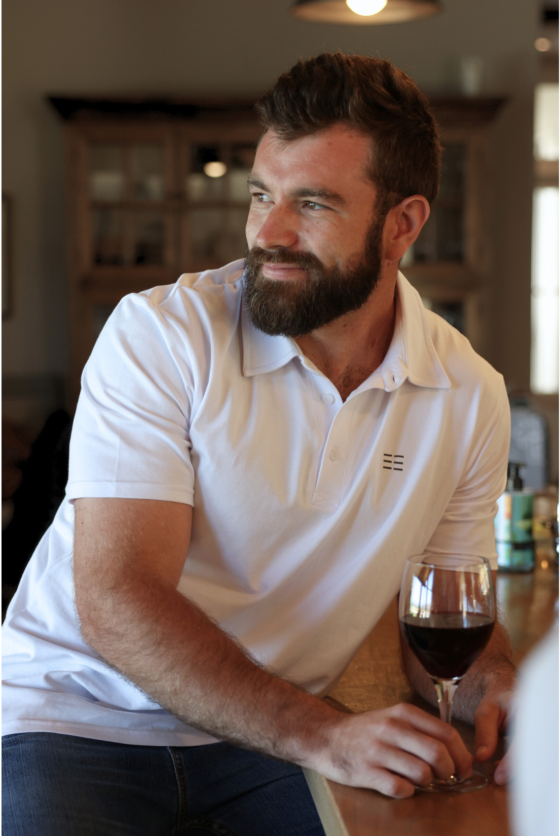 Man sitting at a table with a glass of red wine, wearing a white premium cotton polo shirt.