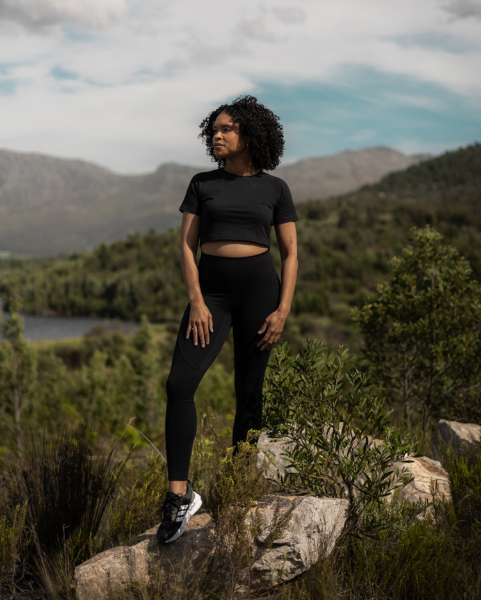 A woman stands outdoors on rocks in a mountainous, grassy landscape wearing a black Breeth branded short-sleeve crop top and black Breeth Holda Ultra leggings, with one black and white sneaker visible.